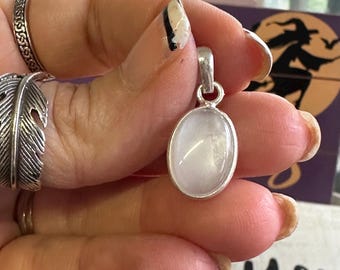 May include: A silver pendant with an oval, translucent, light-colored stone. The pendant is held in a hand with rings and nail polish. The background includes a Halloween-themed decoration with a witch silhouette and the word "you".