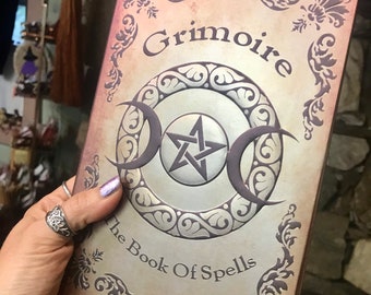 May include: A brown leather-bound book with a silver pentagram and crescent moon design. The book is titled "Grimoire The Book Of Spells".
