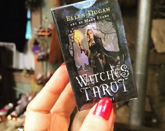 May include: A deck of "Witches Tarot" cards held in a hand with red nail polish. The card box features an illustration of a woman holding a torch, with the text "Ellen Dugan" and "Art by Mark Evans".