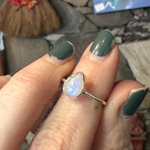 May include: A silver ring with a teardrop-shaped moonstone. The stone is iridescent and has a milky white color.