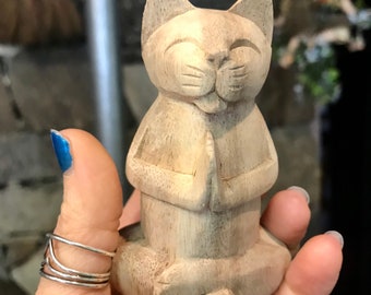 May include: A wooden cat figurine sitting in a meditative pose with its paws folded in front of its chest. The cat has a closed-mouth smile and is carved with detail.