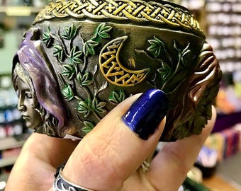 May include: A ceramic incense burner with a Celtic knot design around the top. The burner features a crescent moon with a Celtic knot design, green leaves, and two faces on either side. The faces are partially obscured by the leaves.