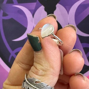 May include: A silver ring with a teardrop-shaped moonstone. The ring is on a finger and the stone is iridescent.