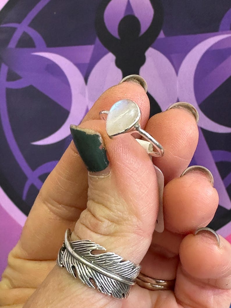 May include: A silver ring with a teardrop-shaped moonstone. The ring is on a finger and the stone is iridescent.