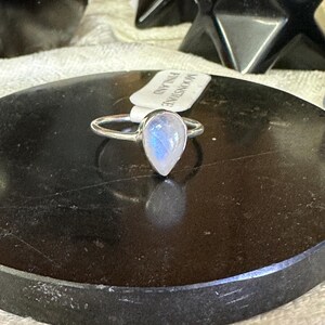 May include: A silver ring with a teardrop-shaped moonstone set in the center. The ring is on a black surface with a tag that reads "Moonstone Finland".