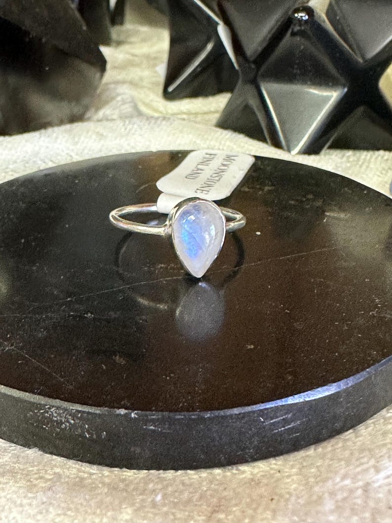 May include: A silver ring with a teardrop-shaped moonstone set in the center. The ring is on a black surface with a tag that reads "Moonstone Finland".