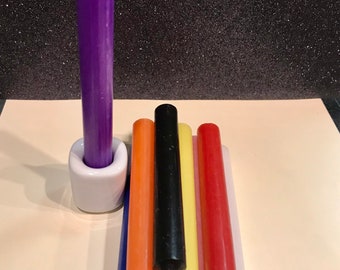 Op de afbeelding: Een verzameling kleurrijke taps toelopende kaarsen. Een lange, paarse kaars staat in een witte keramische houder. Verschillende andere kaarsen in diverse kleuren, waaronder oranje, zwart, geel, rood, roze en wit, zijn in de buurt gerangschikt. De achtergrond is donker en glinsterend.