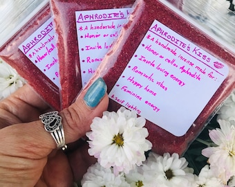 Könnte beinhalten: Drei kleine Beutel mit rotem Räucherwerk, beschriftet mit "Aphrodite's Kiss" mit einem rosa Herzen und "Aphrodite's" mit einem rosa Herzen. Die Beutel sind mit rotem Räucherwerk gefüllt und liegen auf einem Bett aus weißen Blüten.