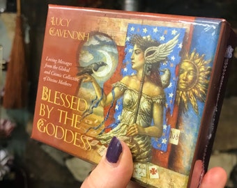 May include: A box of "Blessed by the Goddess" oracle cards by Lucy Cavendish. The box features a colorful illustration of a woman with wings, holding a staff and a raven. The text "Loving Messages from the Global and Cosmic Collective of Divine Mothers" is printed on the box.