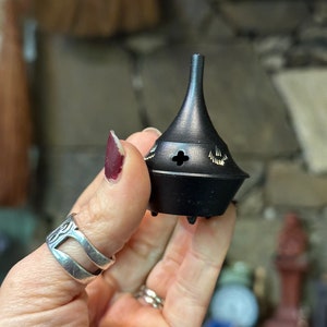 May include: A black metal incense burner with a pointed top and a small hole in the center. The burner has decorative details around the top and bottom.