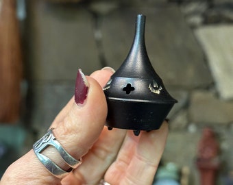 May include: A black metal incense burner with a pointed top and a small hole in the center. The burner has decorative details around the top and bottom.