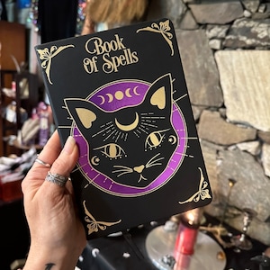Op de afbeelding: Een zwart boek met een gouden en paarse ontwerp met een zwarte kat met gouden ogen en een halve maan. De titel van het boek is "Book of Spells".