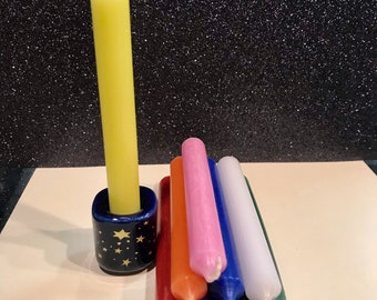 May include: A set of seven small candles in various colors, including red, orange, yellow, green, blue, purple, and white. The candles are arranged in a fan shape and are sitting on a white surface. A larger yellow candle is in a blue and gold star-shaped holder.