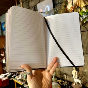Op de afbeelding: Een zwart, gestreept notitieboek met een zwart lint als bladwijzer. Het notitieboek is open op een lege pagina.