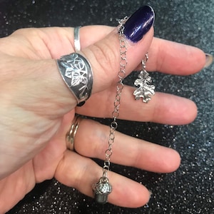 May include: A silver chain necklace with an acorn charm and an oak leaf charm. The necklace is hanging from a person's thumb.