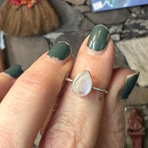 May include: A silver ring with a teardrop-shaped moonstone. The ring is on a finger with green nail polish.