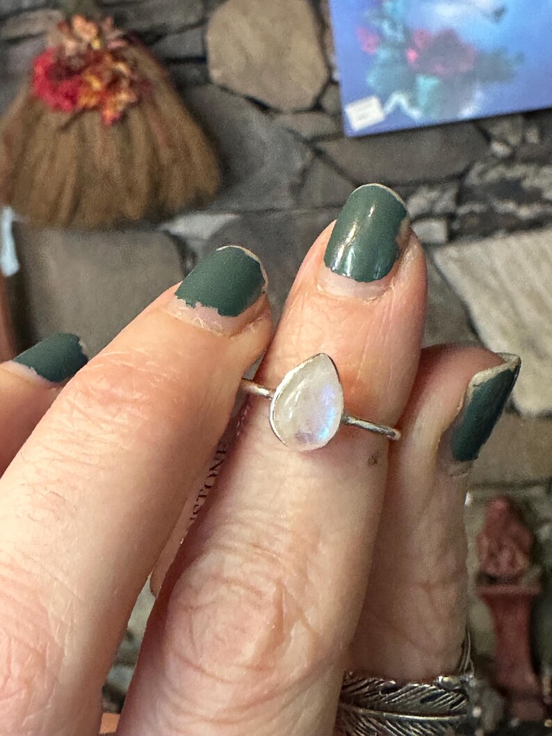 May include: A silver ring with a teardrop-shaped moonstone. The ring is on a finger with green nail polish.