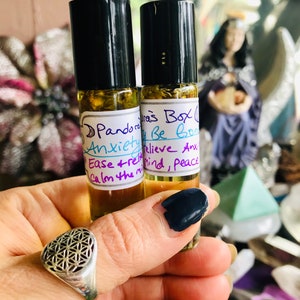 Pode incluir: Dois pequenos frascos de vidro com tampas de rolagem pretas cheios de um líquido amarelo claro. Os frascos são etiquetados com "Pandora's Box", "Anxiety Be Gone", "Believe Anx", "Ease + relieve", "Calm the mind, peace".