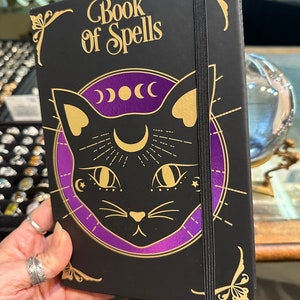 Op de afbeelding: Een zwart notitieboek met een gouden elastiek sluiting en een gouden illustratie van een zwarte kat met een halve maan en sterren op zijn voorhoofd. Het notitieboek heeft de woorden "Book of Spells" in gouden letters op de voorkant.