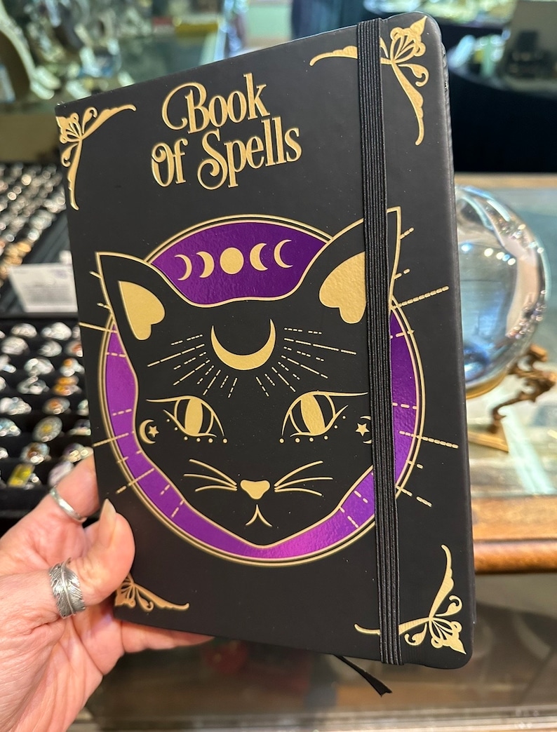 Op de afbeelding: Een zwart notitieboek met een gouden elastiek sluiting en een gouden illustratie van een zwarte kat met een halve maan en sterren op zijn voorhoofd. Het notitieboek heeft de woorden "Book of Spells" in gouden letters op de voorkant.