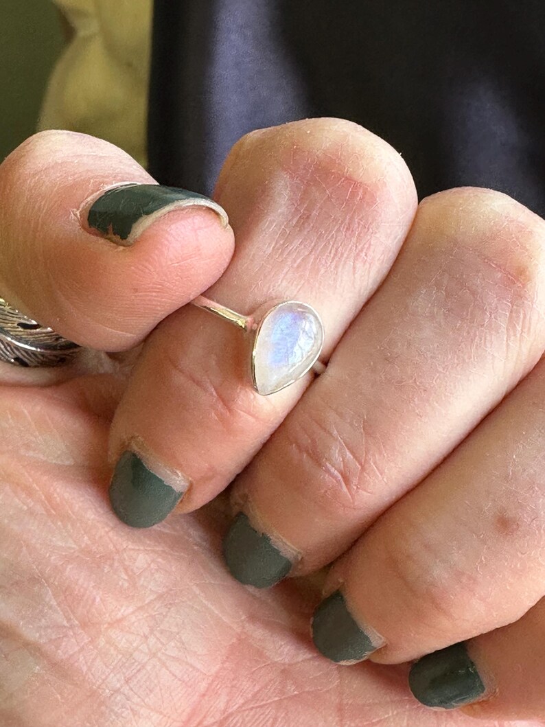 May include: A silver ring with a teardrop-shaped moonstone. The stone is iridescent and has a milky white color.