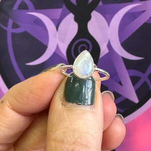 May include: A silver ring with a teardrop-shaped moonstone. The ring is on a finger with green nail polish.