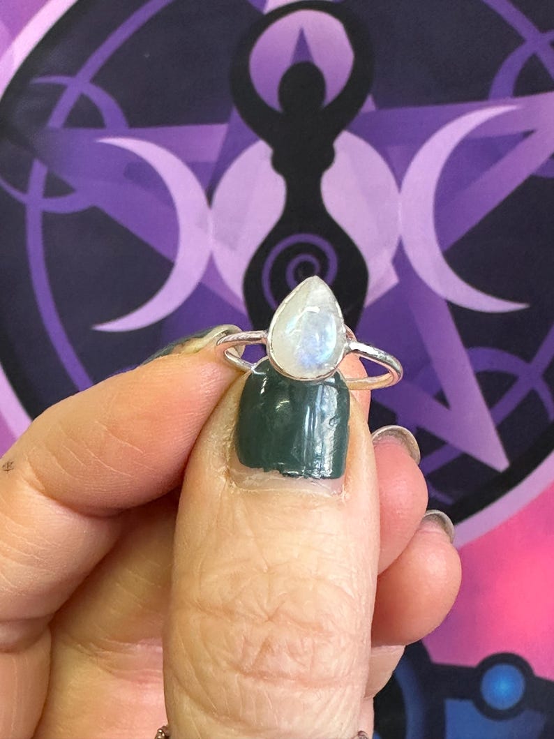 May include: A silver ring with a teardrop-shaped moonstone. The ring is on a finger with green nail polish.