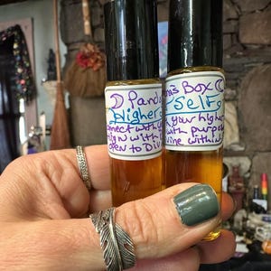 May include: Two glass roll-on bottles with black caps are held in a hand. The bottles have labels with handwritten text: "Pandora's Box Higher Self" and "connect with your higher with purpose open to Divine within". The liquid inside is amber-colored.
