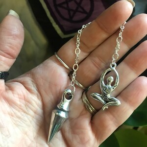 May include: Two silver pendulums, one with a pointed tip and the other with a stylized figure of a woman, hanging from silver chains.