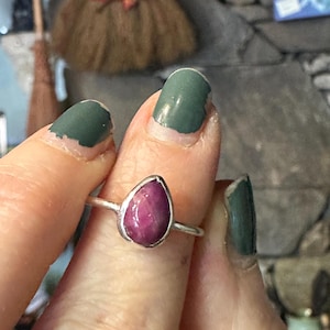 May include: A silver ring with a teardrop-shaped pink gemstone. The ring is on a person's finger.