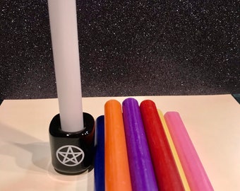 May include: A set of seven small candles in various colors, including blue, green, orange, purple, red, yellow, and pink. The candles are arranged in a pile on a white surface. A larger white candle is in a black candle holder with a white pentagram symbol.