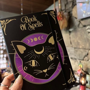 Op de afbeelding: Een zwart notitieboek met een gouden reli&euml;fdesign van een zwarte kat met een halve maan en sterren. Het notitieboek heeft de woorden "Book of Spells" op de voorkant.