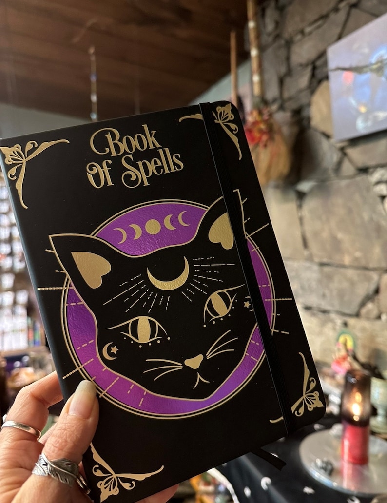 Op de afbeelding: Een zwart notitieboek met een gouden reli&euml;fdesign van een zwarte kat met een halve maan en sterren. Het notitieboek heeft de woorden "Book of Spells" op de voorkant.