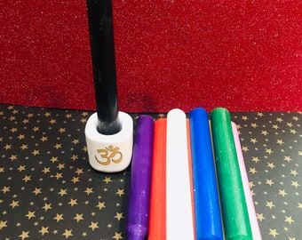 May include: A collection of colorful, slender candles. A black candle stands in a white holder with a gold Om symbol. Additional candles are purple, red, white, blue, green, and pink. The background is red and black with gold stars.