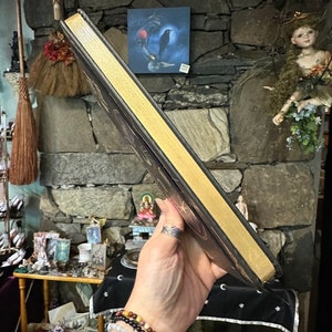 Op de afbeelding: Een zwart lederen gebonden dagboek met goudkleurige randen en een decoratief ontwerp op de omslag. Het dagboek wordt vastgehouden in een hand met een zilveren ring aan de wijsvinger.