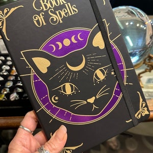 Op de afbeelding: Een zwart notitieboek met een gouden elastiek sluiting en een gouden en paarse illustratie van een kat met een halve maan en sterren. De omslag van het notitieboek staat "Book of Spells".