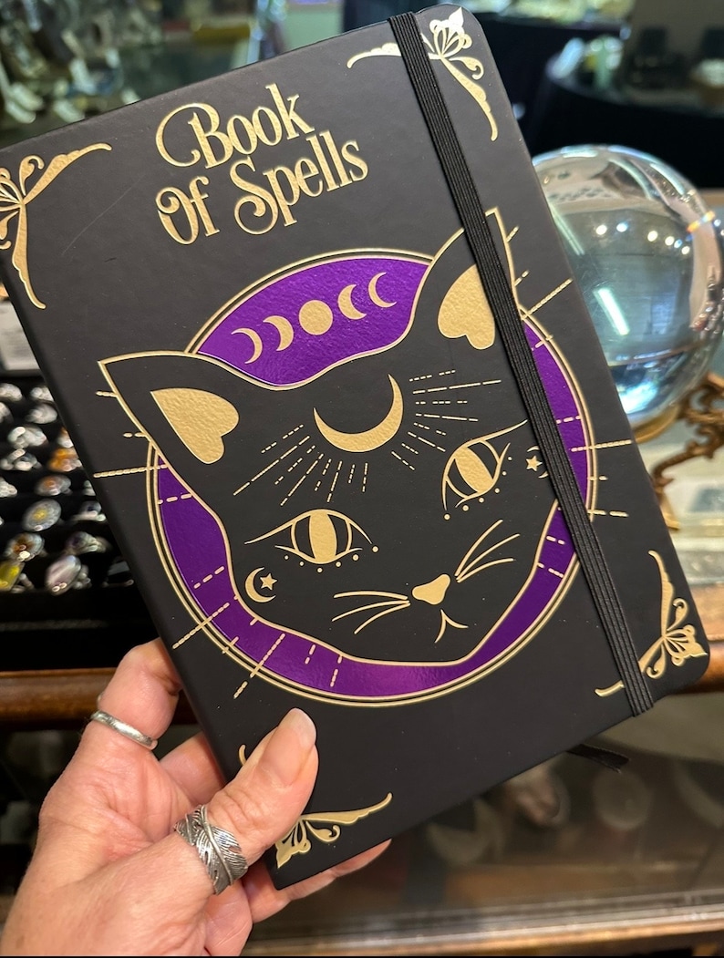 Op de afbeelding: Een zwart notitieboek met een gouden elastiek sluiting en een gouden en paarse illustratie van een kat met een halve maan en sterren. De omslag van het notitieboek staat "Book of Spells".