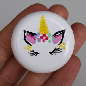 Może przedstawiać: Okrągła, biała ozdoba ceramiczna z namalowaną twarzą jednorożca. Jednorożec ma żółty róg, różowe uszy, czarne rzęsy i wianek z żółtych, różowych, fioletowych i zielonych kwiatów.