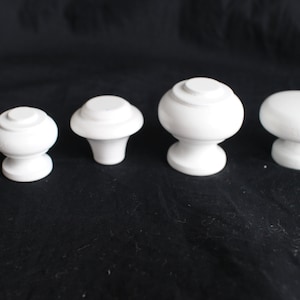 Może przedstawiać: Pięć białych ceramicznych gałek do szafek w różnych rozmiarach. Gałki mają zaokrąglony kształt przypominający grzyb z podniesionym, ozdobnym rantem.