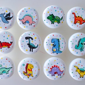 Może przedstawiać: Dwunastu białych, okrągłych gałek z kolorowymi wzorami dinozaurów. Każda gałka ma innego dinozaura na kolorowym tle w kropki.