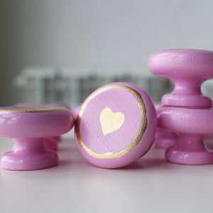 May include: A group of pink wooden drawer knobs with gold accents. Some of the knobs have a gold heart design.