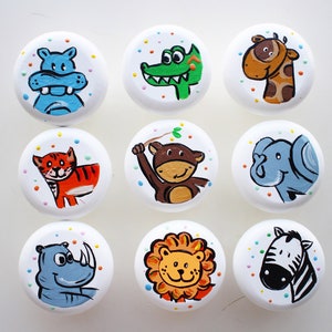 以下が含まれることがあります： カラフルな動物のイラストが描かれた白い丸いノブが9つあります。動物は、カバ、ワニ、キリン、トラ、サル、ゾウ、サイ、ライオン、シマウマです。各ノブは、カラフルなコンフェッティが散りばめられた白い背景になっています。