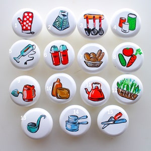 May include: Fifteen white round ceramic knobs with painted kitchen-themed images. The images include a red oven mitt, a green grater, a set of kitchen utensils, a can of beans, a hand mixer, salt and pepper shakers, a loaf of bread, a tomato and cucumber, a red mixer, a bottle of oil, a red kettle, a bowl of herbs, a ladle, measuring cups, and a pair of kitchen shears.