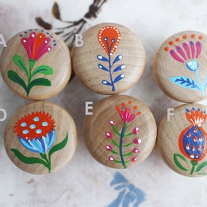 Può includere: Sei pomelli in legno con disegni floreali dipinti a mano. Ogni pomello presenta un fiore diverso con uno schema di colori unico. I fiori sono dipinti in tonalità di rosso, arancione, blu, verde e bianco.