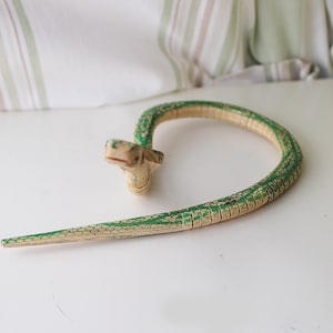 Puede incluir: Una serpiente de juguete de madera pintada de verde y beige. La serpiente est&aacute; enroscada en forma de c&iacute;rculo.
