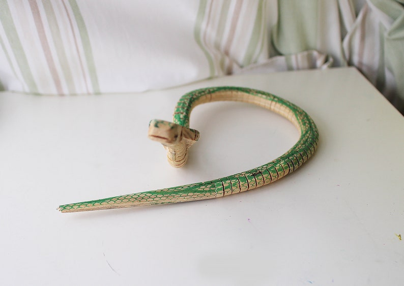 Puede incluir: Una serpiente de juguete de madera pintada de verde y beige. La serpiente est&aacute; enroscada en forma de c&iacute;rculo.