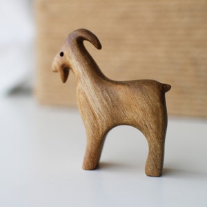 May include: A handcrafted wooden goat figurine. The goat is carved from light brown wood, with visible grain. It has a curved horn, a small beard, and stands on four legs. The figurine is a decorative item.