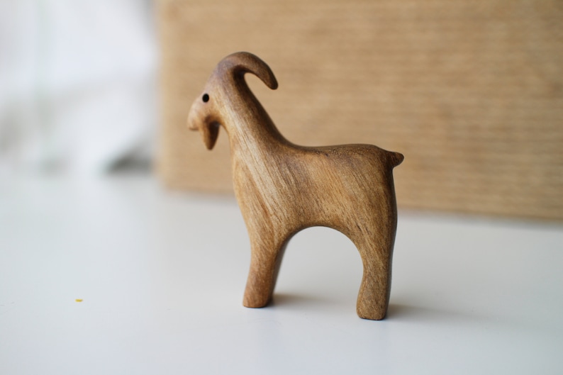 May include: A handcrafted wooden goat figurine. The goat is carved from light brown wood, with visible grain. It has a curved horn, a small beard, and stands on four legs. The figurine is a decorative item.