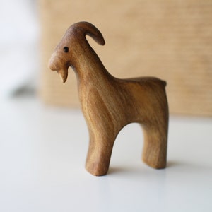 May include: A handcrafted wooden goat figurine. The goat is carved from a light brown wood, with visible grain. It has a curved horn, a small beard, and a simple, stylized form. The figurine stands on four legs, with a smooth, polished finish.