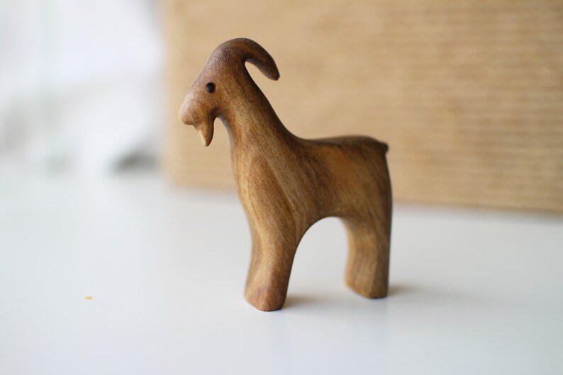 May include: A handcrafted wooden goat figurine. The goat is carved from a light brown wood, with visible grain. It has a curved horn, a small beard, and a simple, stylized form. The figurine stands on four legs, with a smooth, polished finish.